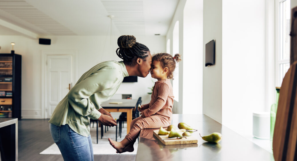 Mom leaning in to kiss her toddler daughter who is sitting on the counter next to sliced fruit on a cutting board