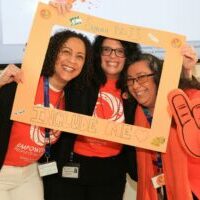 3 women wearing orange, smiling while holding a hand-made Inclusion Day photo frame for Summit DD's Inclusion Day